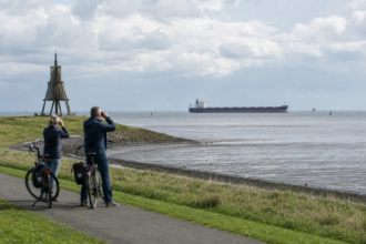 Radreisende beobachten an der Kugelbake in Cuxhaven ein Schiff auf der Elbe [Bildinhalt mit Ki bearbeitet]