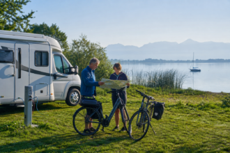 Wohnmobilreisende planen an einem Chiemsee-Campingplatz eine Fahrradtour vor dem Alpenpanorama [Bildinhalt mit Ki bearbeitet]